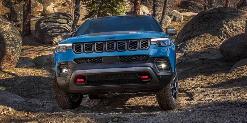 Blue Jeep Compass Trailhawk SUV facing forward on a rocky dirt road with trees and large boulders