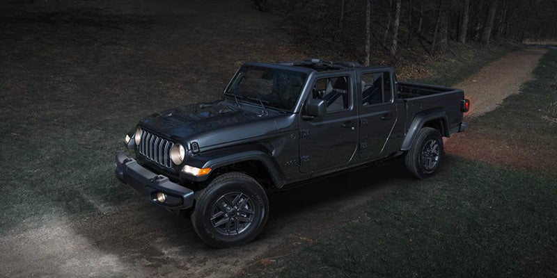 Dark gray Jeep Gladiator truck parked on a dirt path in a dark, wooded area with its headlights on