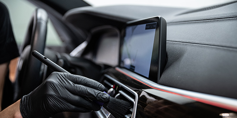A person in a black glove uses a detailing brush to clean a car's dashboard near the vents and display screen