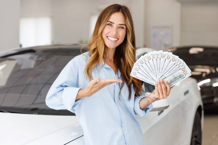 Smiling woman holding fanned $100 bills, gesturing, next to a white car in a showroom