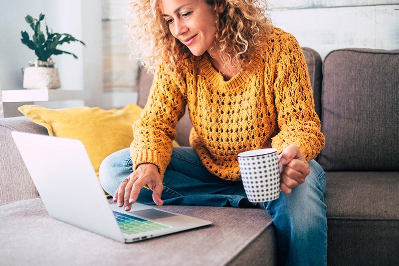 Woman with curly hair in a cozy sweater, sitting on a sofa, smiling while typing on a laptop and holding a mug