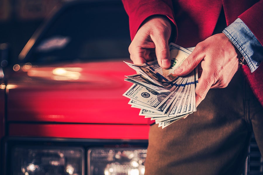 Person holding a large fan of hundred-dollar bills in front of a blurred red car